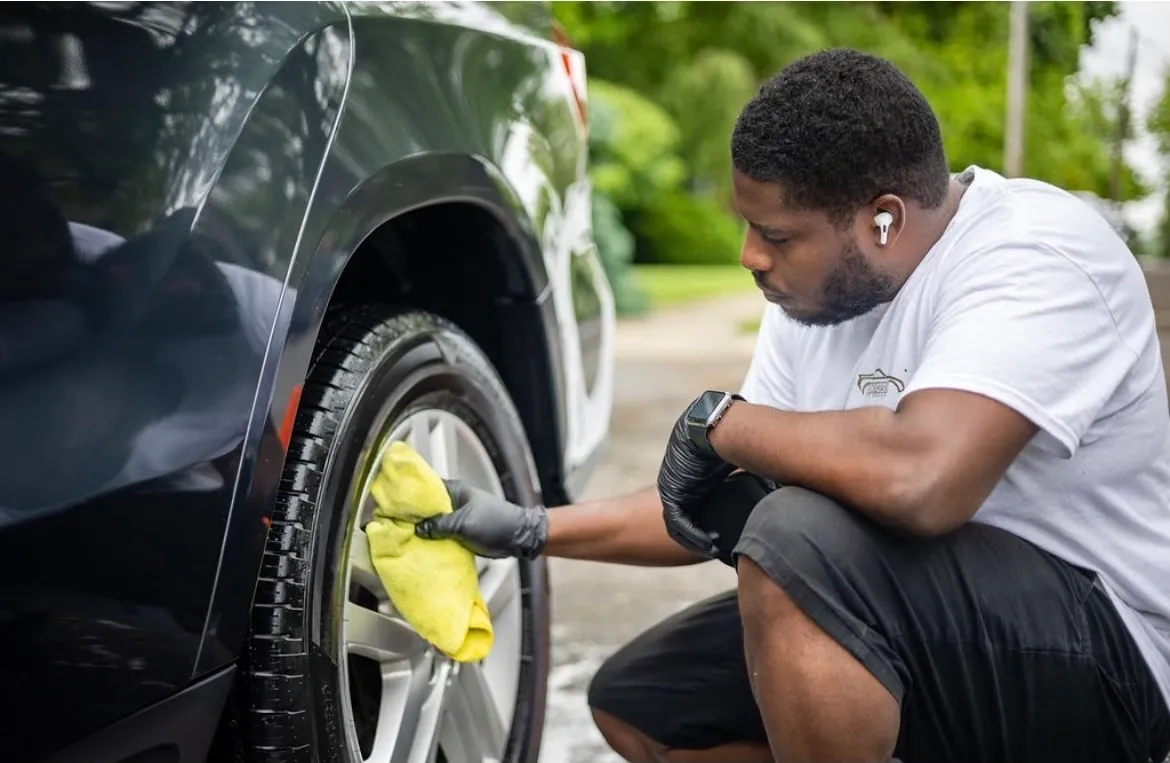 A focused individual meticulously performing auto detailing on a car's wheel with a yellow cloth while wearing earphones and protective gloves.
