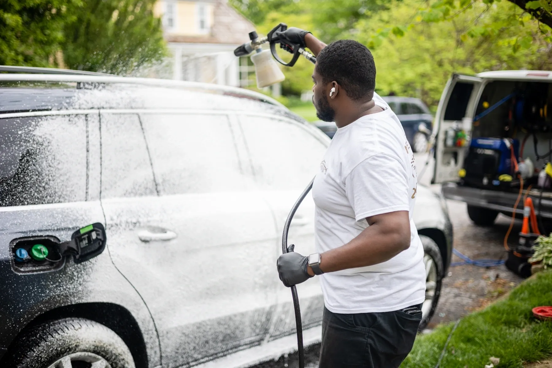 A man diligently performing auto detailing on a car with a high-pressure washer in a residential driveway.
