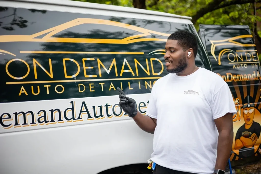 A smiling person stands next to a van adorned with 'auto detailing' graphics, holding a key or device, possibly ready to start a day of work.