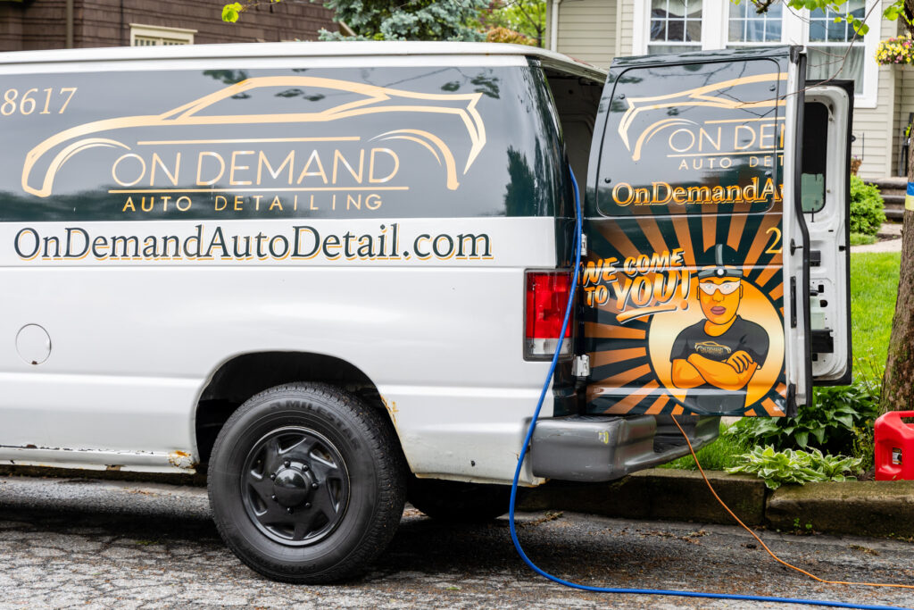 A white van with "On Demand Auto Detailing" branding is parked on a Leander street. The back door is open, showcasing a cartoon character and the text, "We come to you!" Cables extend into the van as nearby houses and trees provide a charming backdrop.