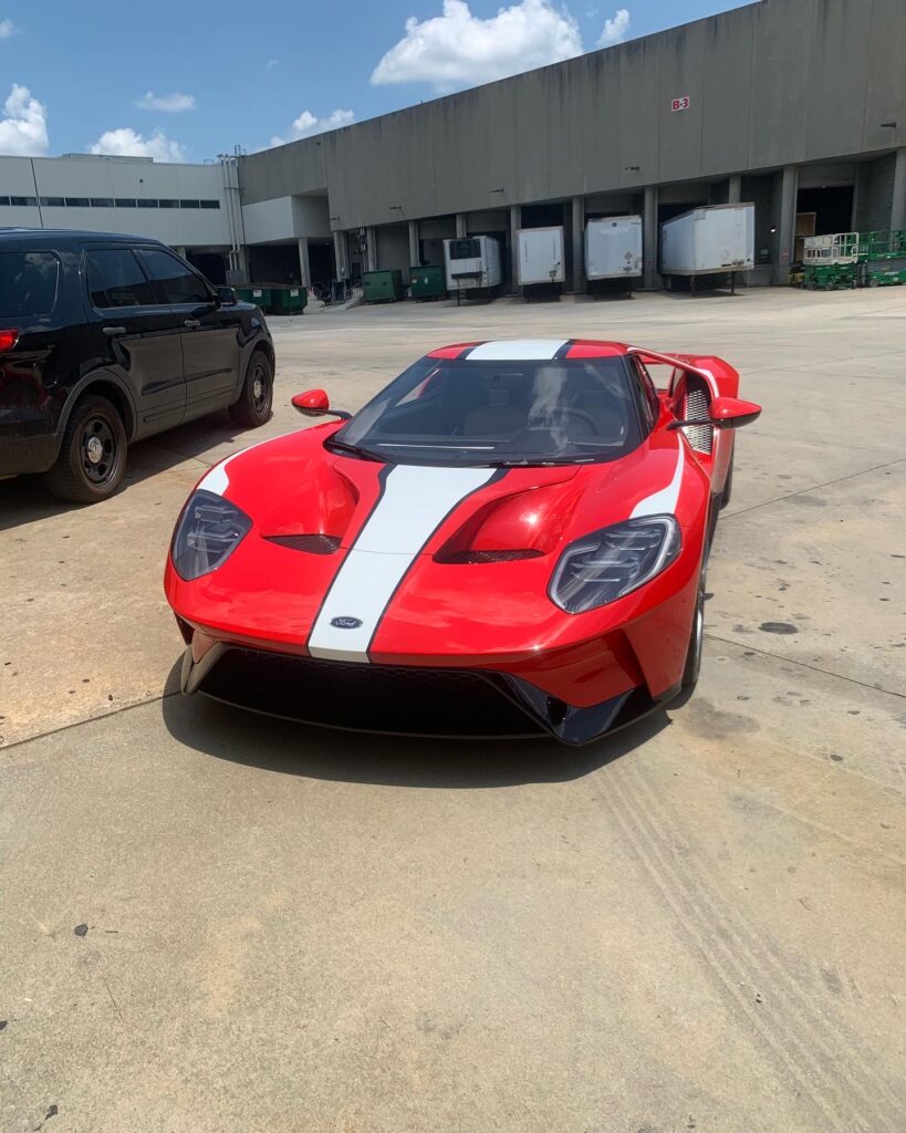 A red sports car with a white racing stripe gleams from expert car detailing as it sits on the concrete surface. Set against a backdrop of loading docks at a large building in Leander, beneath a blue sky dotted with clouds, another black vehicle rests nearby.