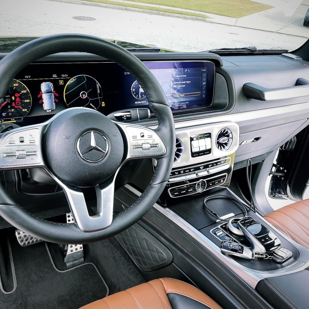 The interior of a luxury car boasts a meticulously detailed design, with a Mercedes steering wheel, digital dashboard, and infotainment screen. The cabin is sleek with brown leather seats, silver accents, and a gear console. Sunlight illuminates Leander's pristine automotive artistry.