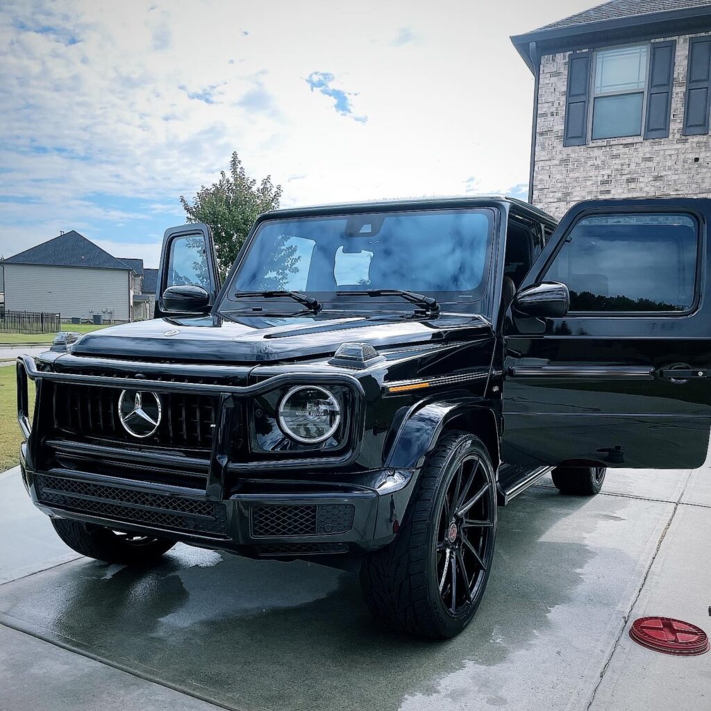 A black Mercedes-Benz G-Class SUV gleams on a Leander driveway, its front passenger door invitingly open. The vehicle, expertly pampered with car detailing, shines against the backdrop of a quiet residential neighborhood and a partly cloudy sky.